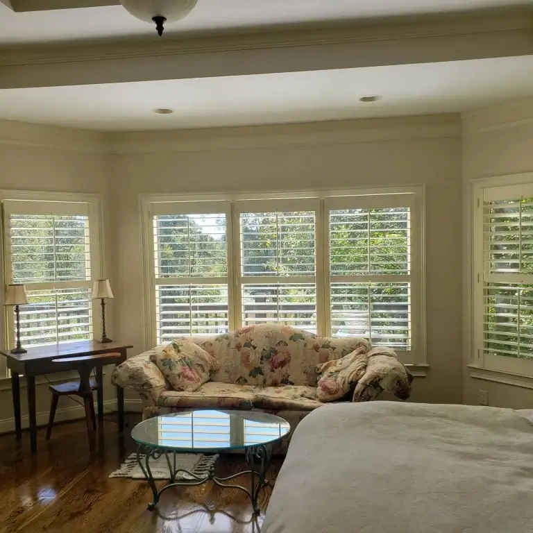 Bright living area with a floral-patterned sofa in front of large bay windows with white shutters, a glass-topped coffee table, a dark wood desk with two lamps on the left, and a bed partially visible in the foreground.