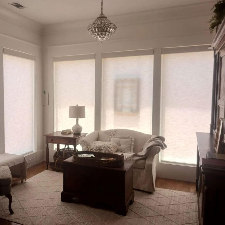 Bright living room with three large windows, a crystal chandelier, and a white sofa with pillows. a wooden coffee table sits in front of the sofa.